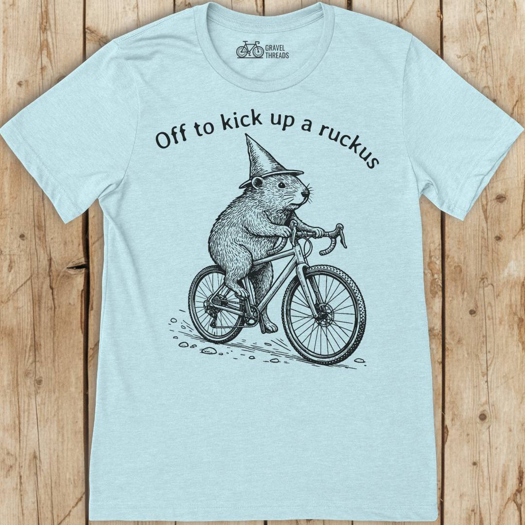 Light blue t-shirt with a graphic of a beaver on a bicycle and text 'Off to kick up a ruckus' on a wooden surface.