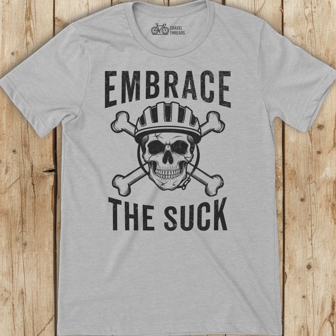 Gray t-shirt with skull and crossbones graphic and text 'Embrace The Suck' on a wooden background