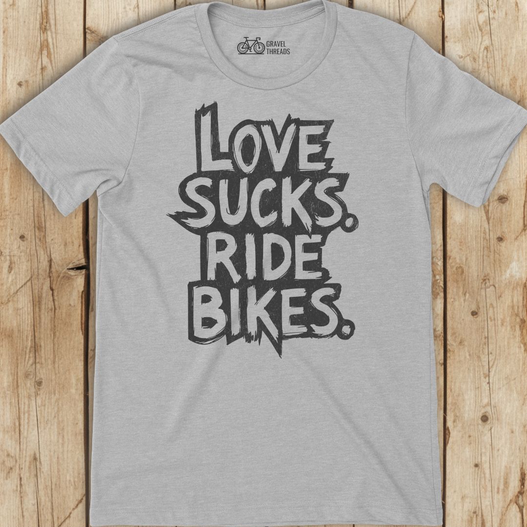 Gray t-shirt with 'I Love Sucks. Ride Bikes.' text on a wooden surface