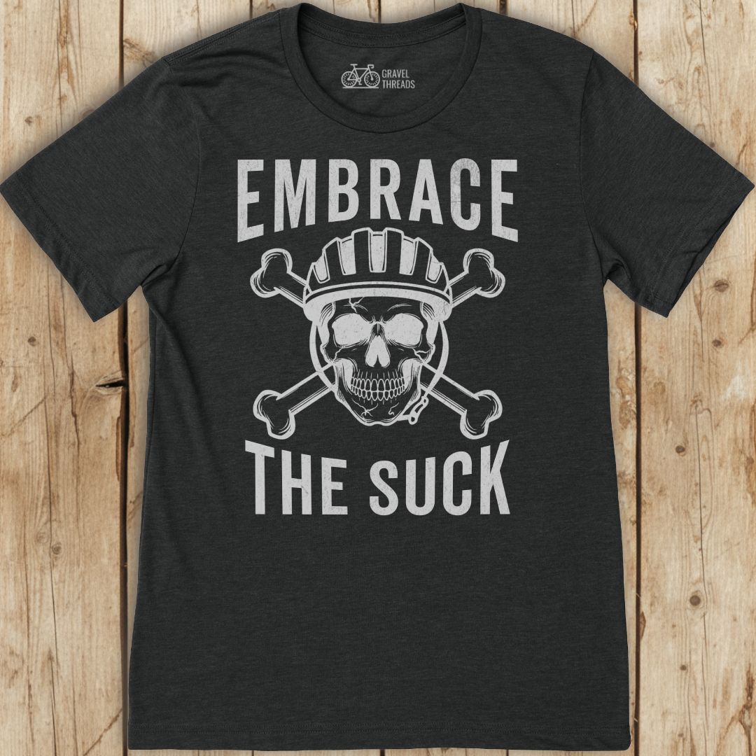 Black t-shirt with skull and crossbones design and text 'Embrace The Suck' on a wooden background
