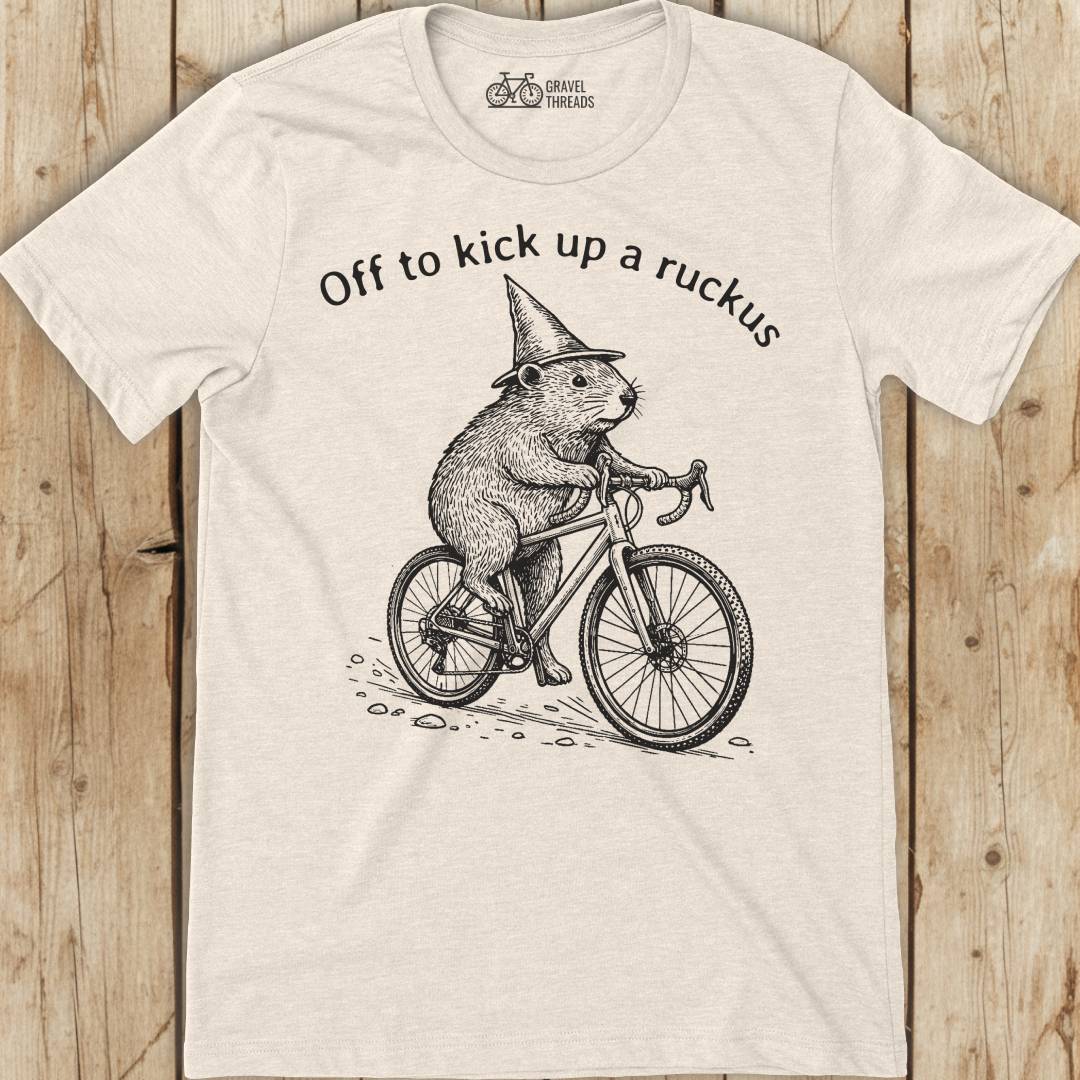 White t-shirt with a graphic of a beaver riding a bicycle and text 'Off to kick up a ruckus' on a wooden surface.