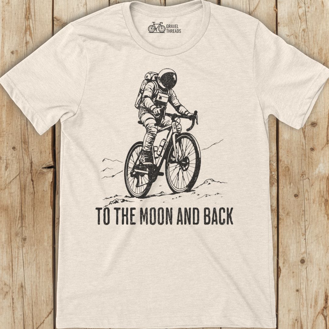 Beige t-shirt with an astronaut riding a bike graphic and 'To the Moon and Back' text on a wooden surface.