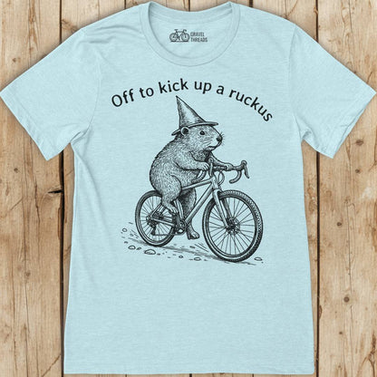 Light blue t-shirt with a graphic of a beaver on a bicycle and text 'Off to kick up a ruckus' on a wooden surface.