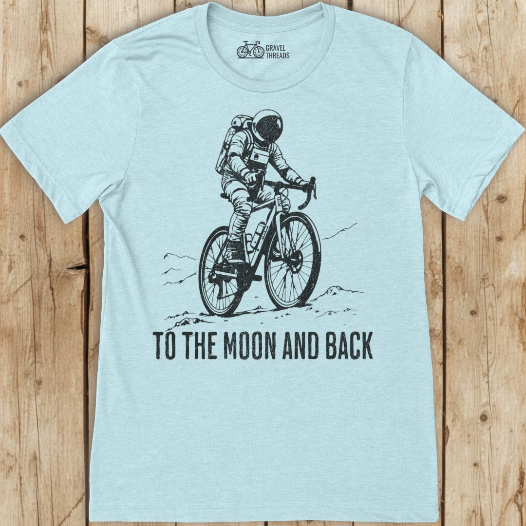 Light blue t-shirt with an astronaut riding a bike graphic and 'To the Moon and Back' text on a wooden surface.