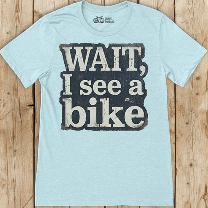 Light blue t-shirt with 'Wait, I see a bike' text on a wooden surface