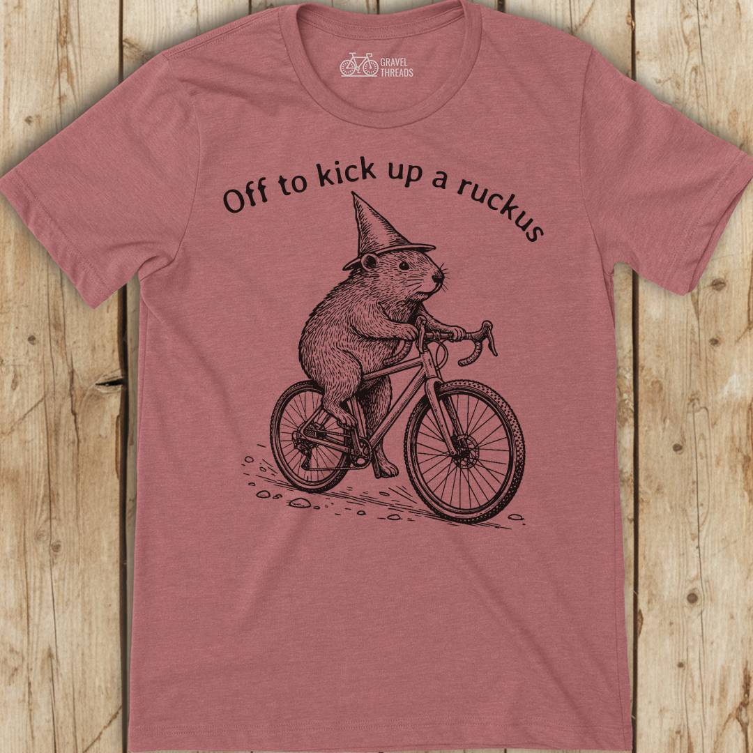 Red t-shirt with a graphic of a beaver on a bicycle and text 'Off to kick up a ruckus' on a wooden surface.