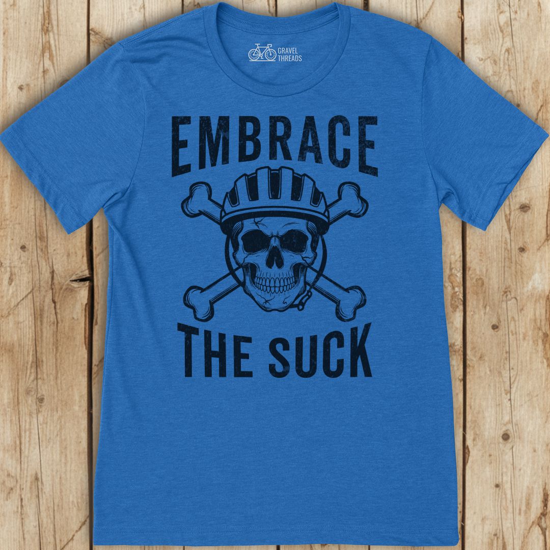 Blue t-shirt with skull graphic and text 'Embrace The Suck' on a wooden surface