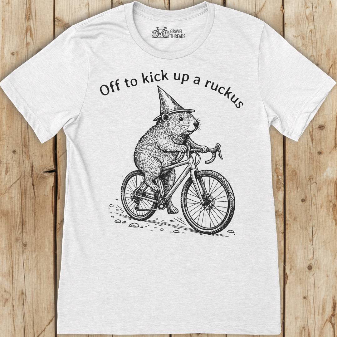 White t-shirt with a graphic of a beaver on a bicycle and text 'Off to kick up a ruckus' on a wooden surface.