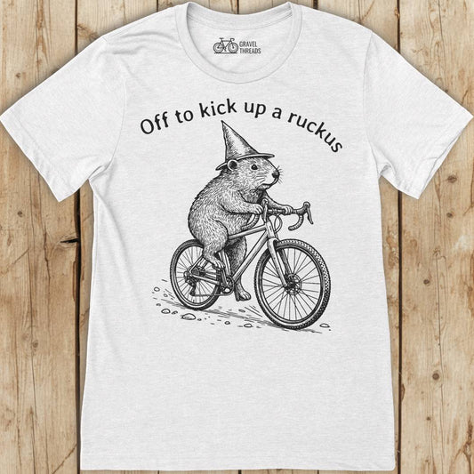 White t-shirt with a graphic of a beaver on a bicycle and text 'Off to kick up a ruckus' on a wooden surface.